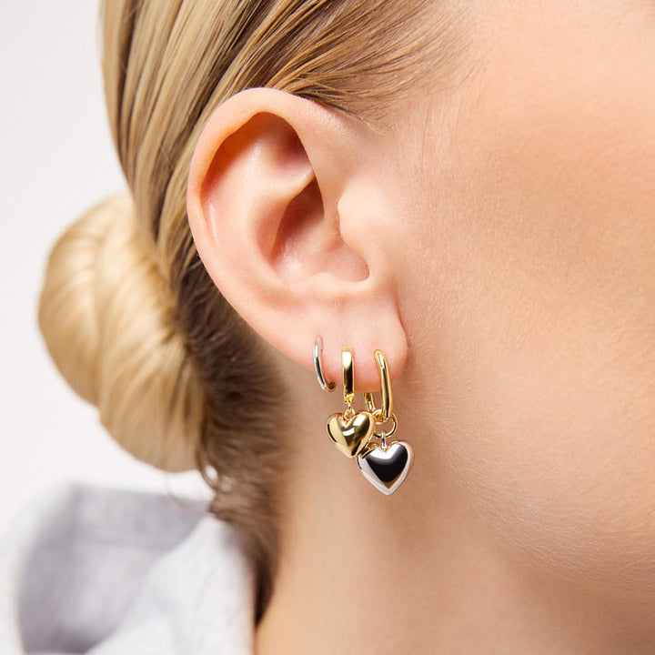 Gold hoop earrings with heart charms worn by a person with blonde hair.