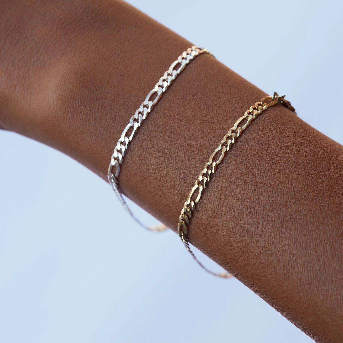 Two gold-toned Figaro chain bracelets worn on a person's wrist against a light blue background.