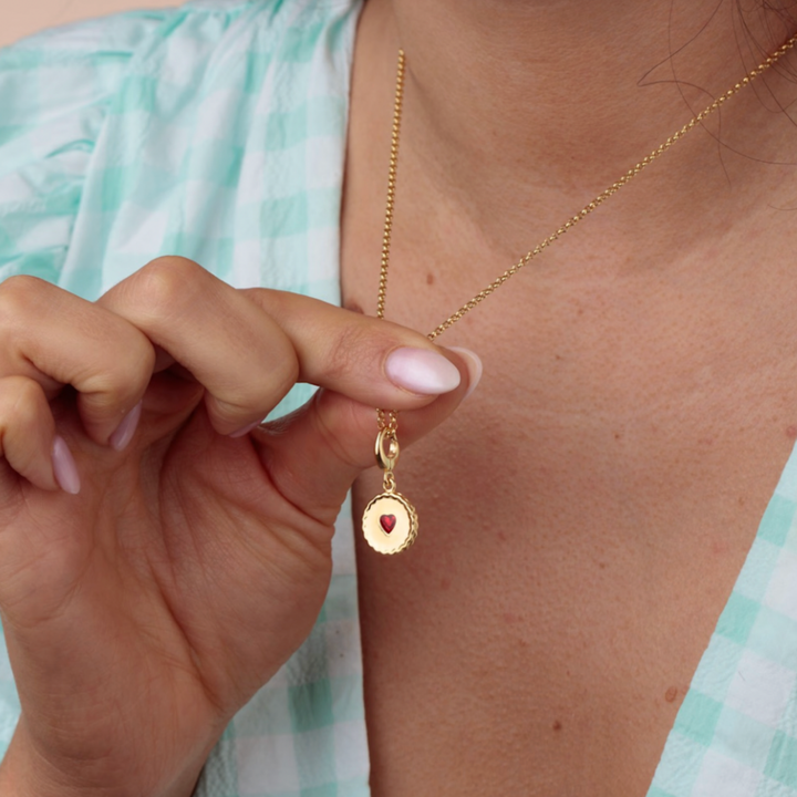 Gold necklace with a red heart pendant held by a hand against a light green background