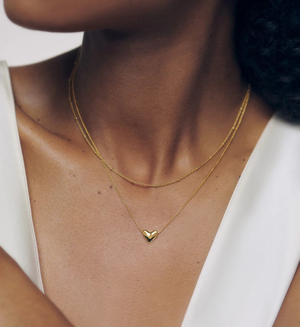 A close-up of a model wearing a delicate gold double-layer necklace with a small heart-shaped pendant on a white top.