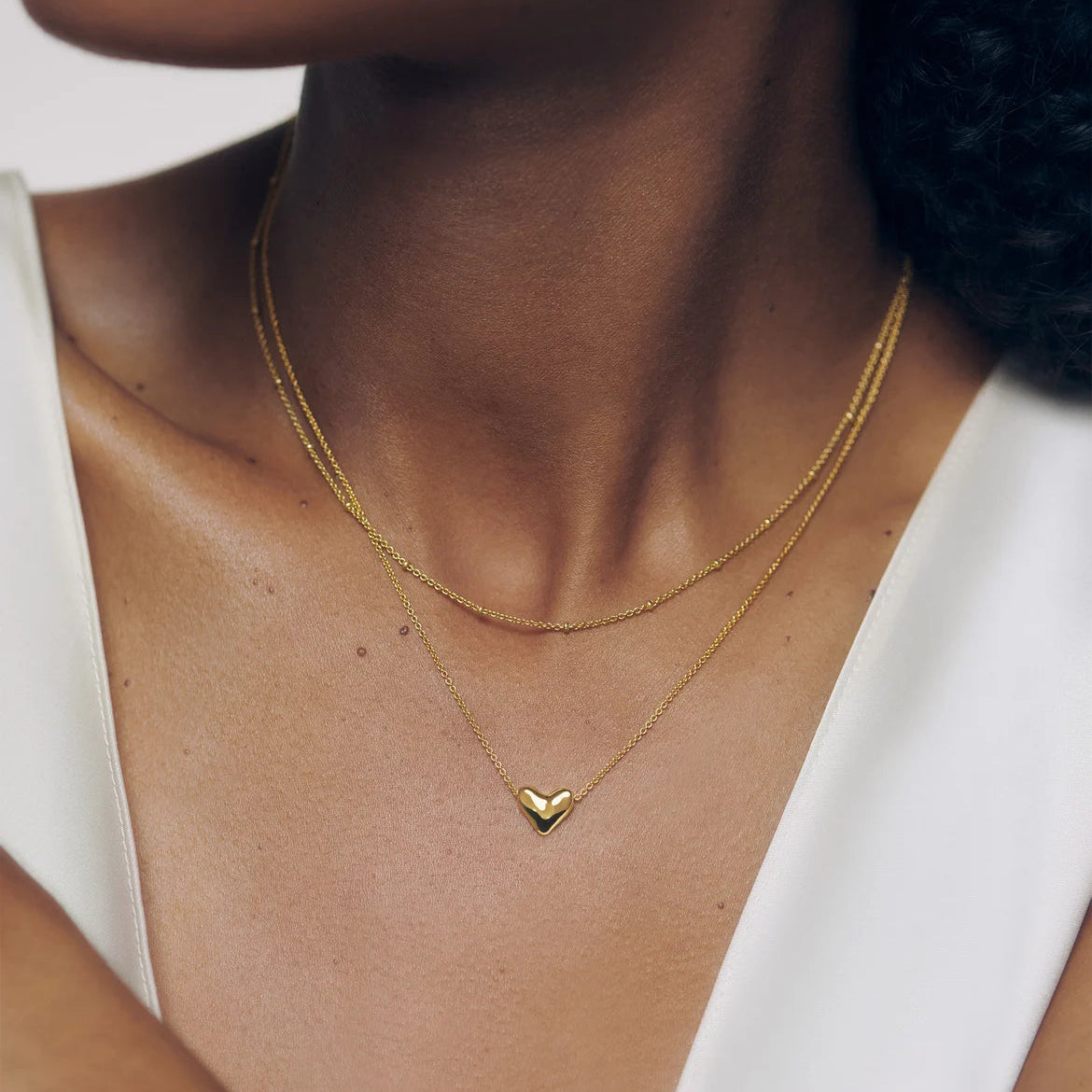 A close-up of a model wearing a delicate gold double-layer necklace with a small heart-shaped pendant on a white top.