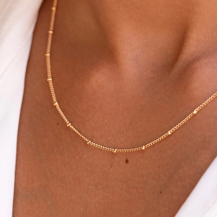 A delicate rose gold chain necklace with small bead-like links, worn on a model's neck over a white top.