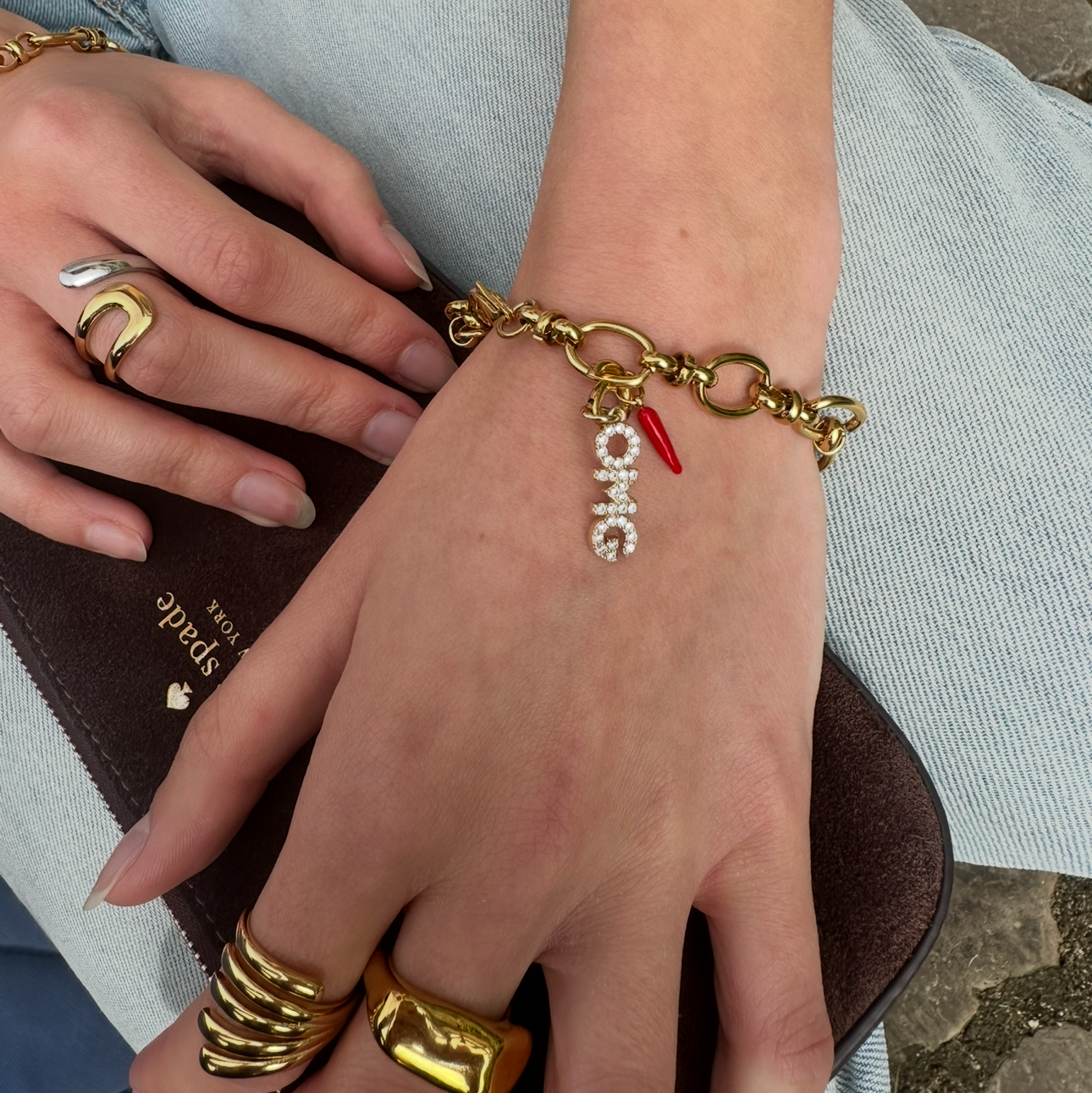Close-up of a hand wearing a gold bracelet with charms, holding a brown clutch.