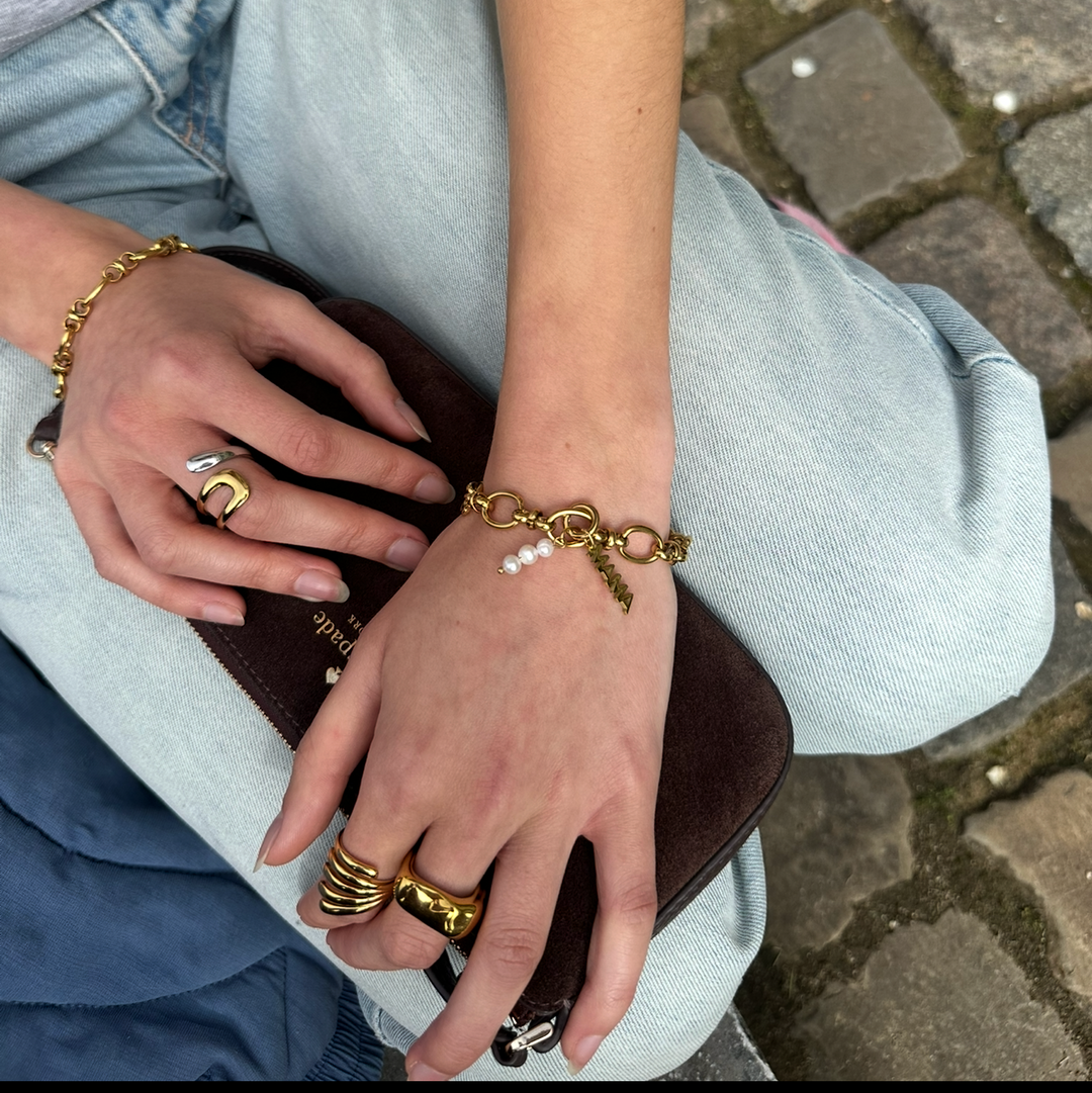 Close-up of hands with gold jewelry holding a brown clutch on a stone pavement.