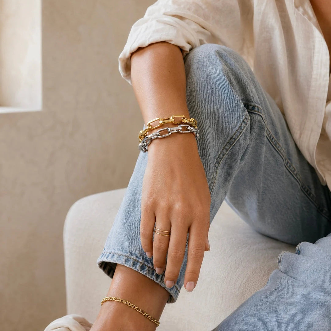A close-up lifestyle shot of a person wearing a stack of three chunky chain bracelets on their wrist, including one gold chain, one silver chain, and one gold link bracelet, paired with light blue jeans and a cream shirt.