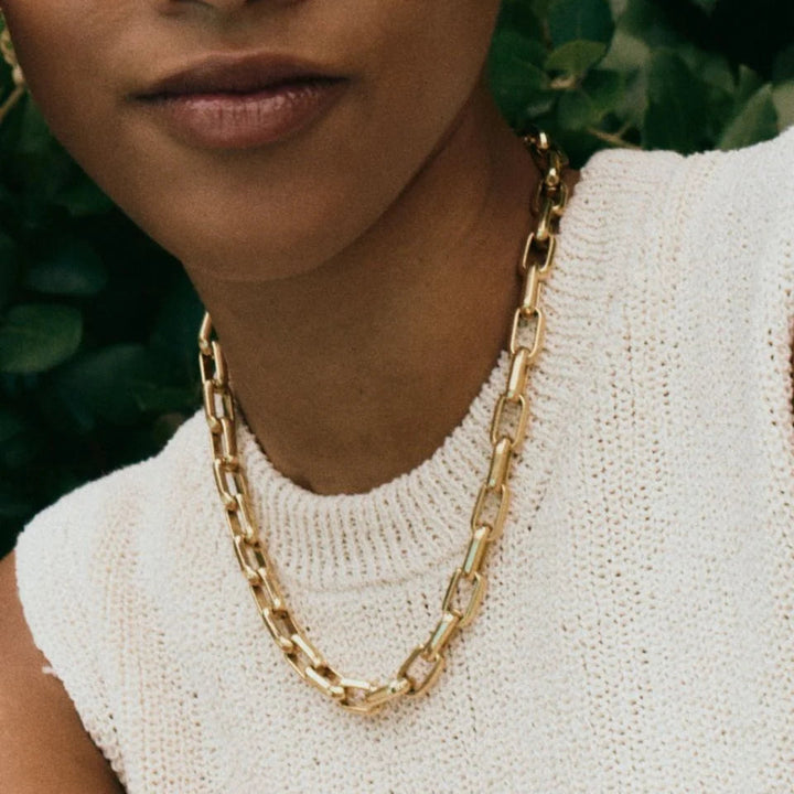 A close-up of a model wearing a chunky gold chain necklace with large links, paired with a white knit top and gold hoop earrings, set against a soft-focus background of green foliage.