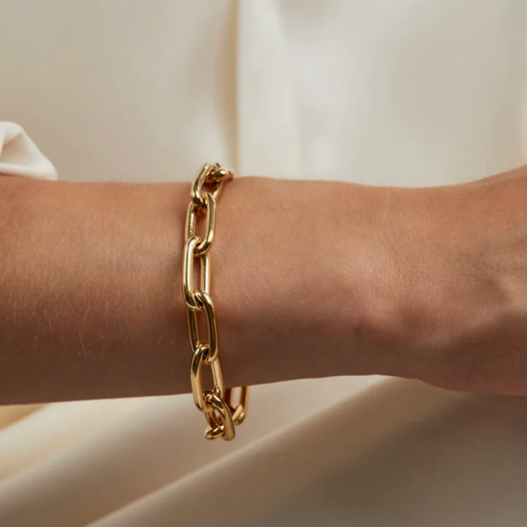 A close-up of a person's wrist wearing a thick, polished gold chain link bracelet against a soft, draped cream-colored fabric background.