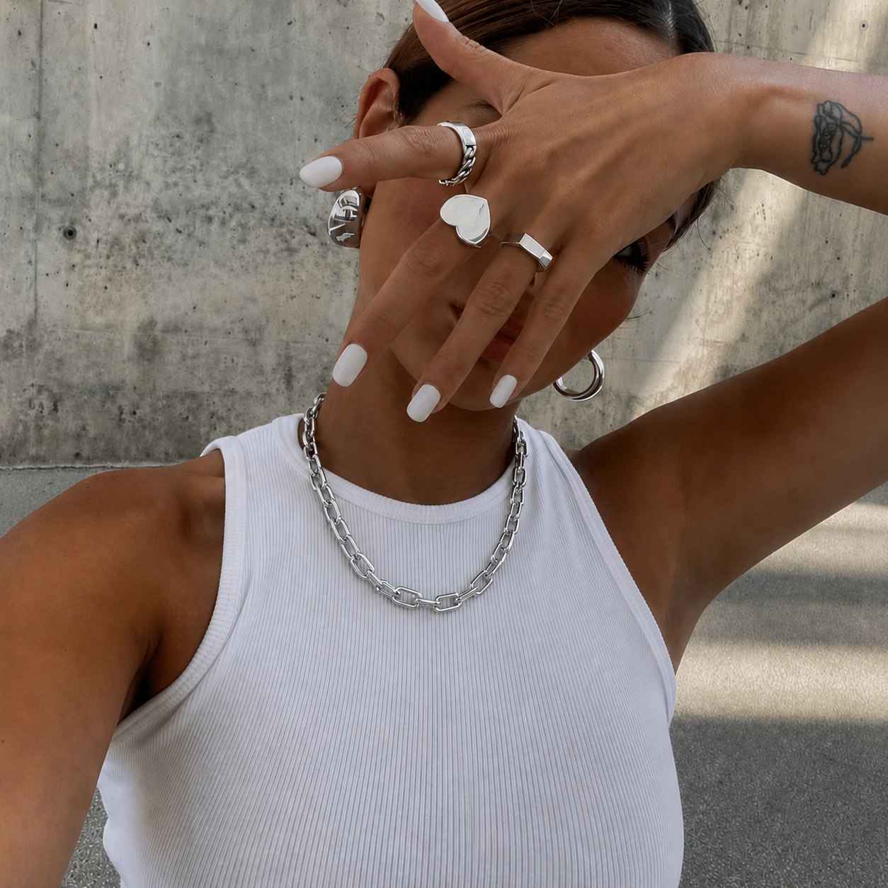 A close-up lifestyle shot of a person wearing a chunky silver chain necklace, silver rings, and silver hoop earrings, wearing a white ribbed tank top against a concrete wall.