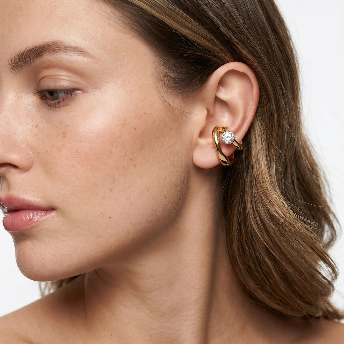Close-up of a woman wearing a gold earring with a clear gemstone on a neutral background