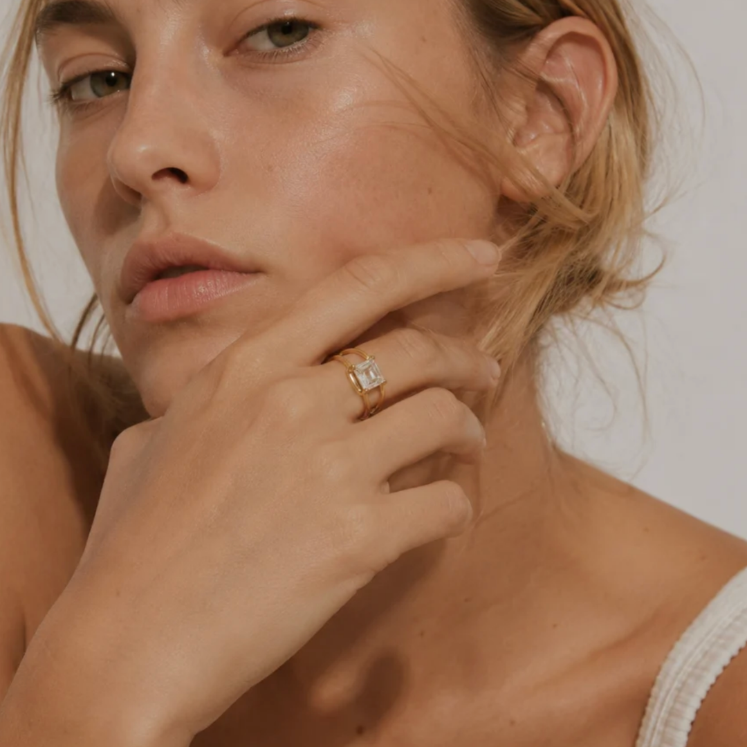 A close-up lifestyle shot of a woman wearing a gold ring with a rectangular clear gemstone on her finger, set against a warm neutral background.