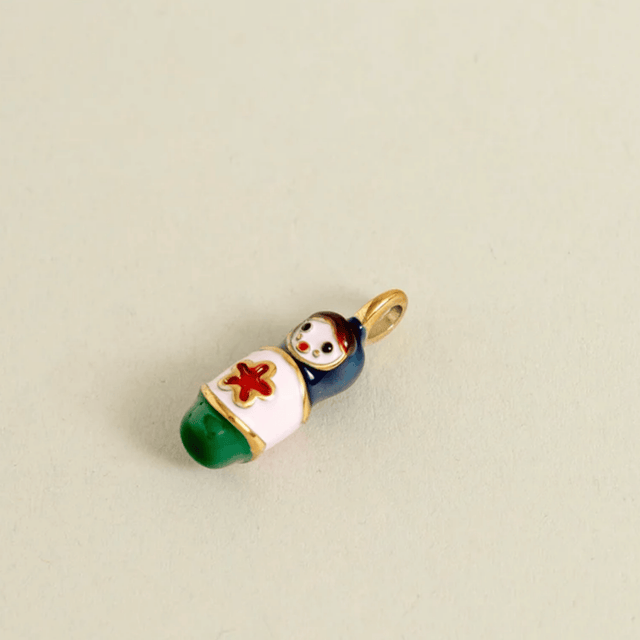 A small enamel charm shaped like a baby doll wrapped in a white blanket with a red cross, green feet, and a dark blue body, attached to a gold ring loop, shown on a plain beige background.
