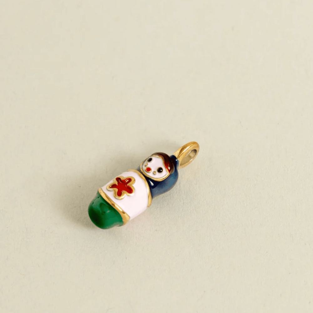 A small enamel charm shaped like a baby doll wrapped in a white blanket with a red cross, green feet, and a dark blue body, attached to a gold ring loop, shown on a plain beige background.