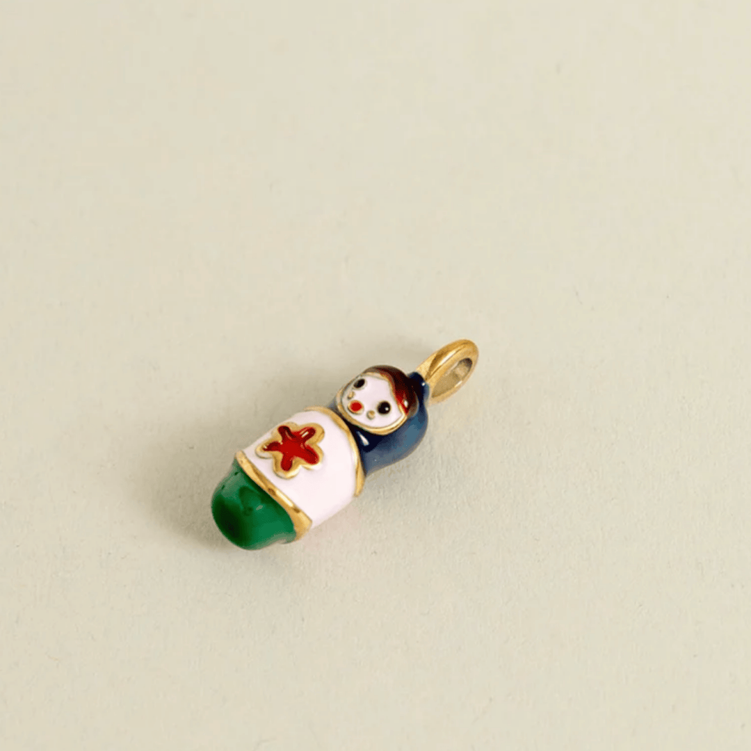 A small enamel charm shaped like a baby doll wrapped in a white blanket with a red cross, green feet, and a dark blue body, attached to a gold ring loop, shown on a plain beige background.