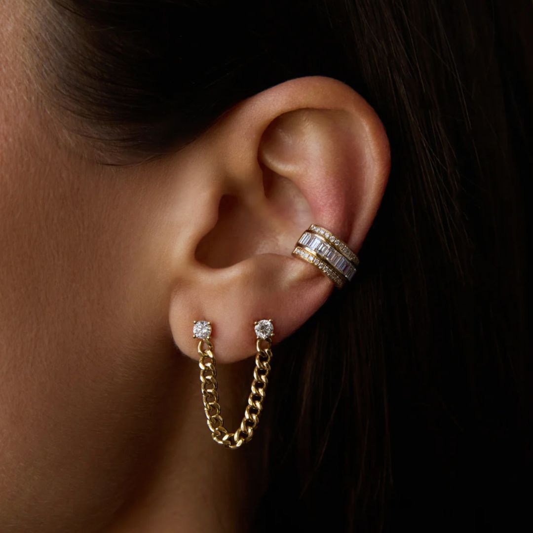 Close-up of an ear with gold chain earring and diamond stud earrings, wearing a silver ring.