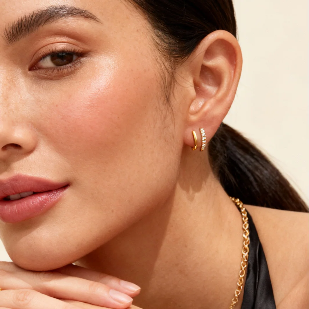 Close-up of a woman wearing gold hoop earrings and a necklace against a neutral background