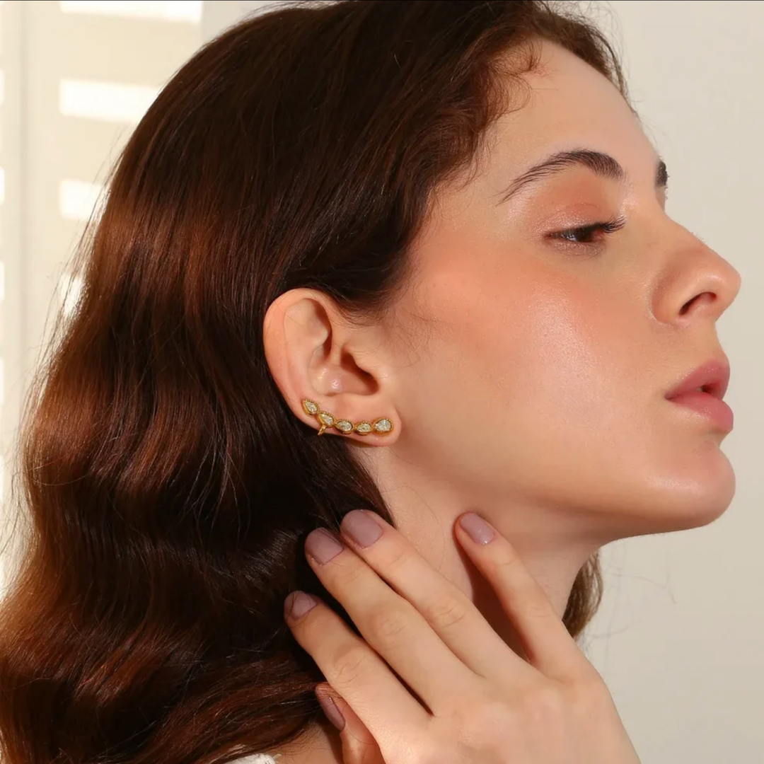 A close-up of a woman with wavy brown hair wearing a delicate gold ear cuff adorned with small green and white stones, with her hand gently touching her neck.