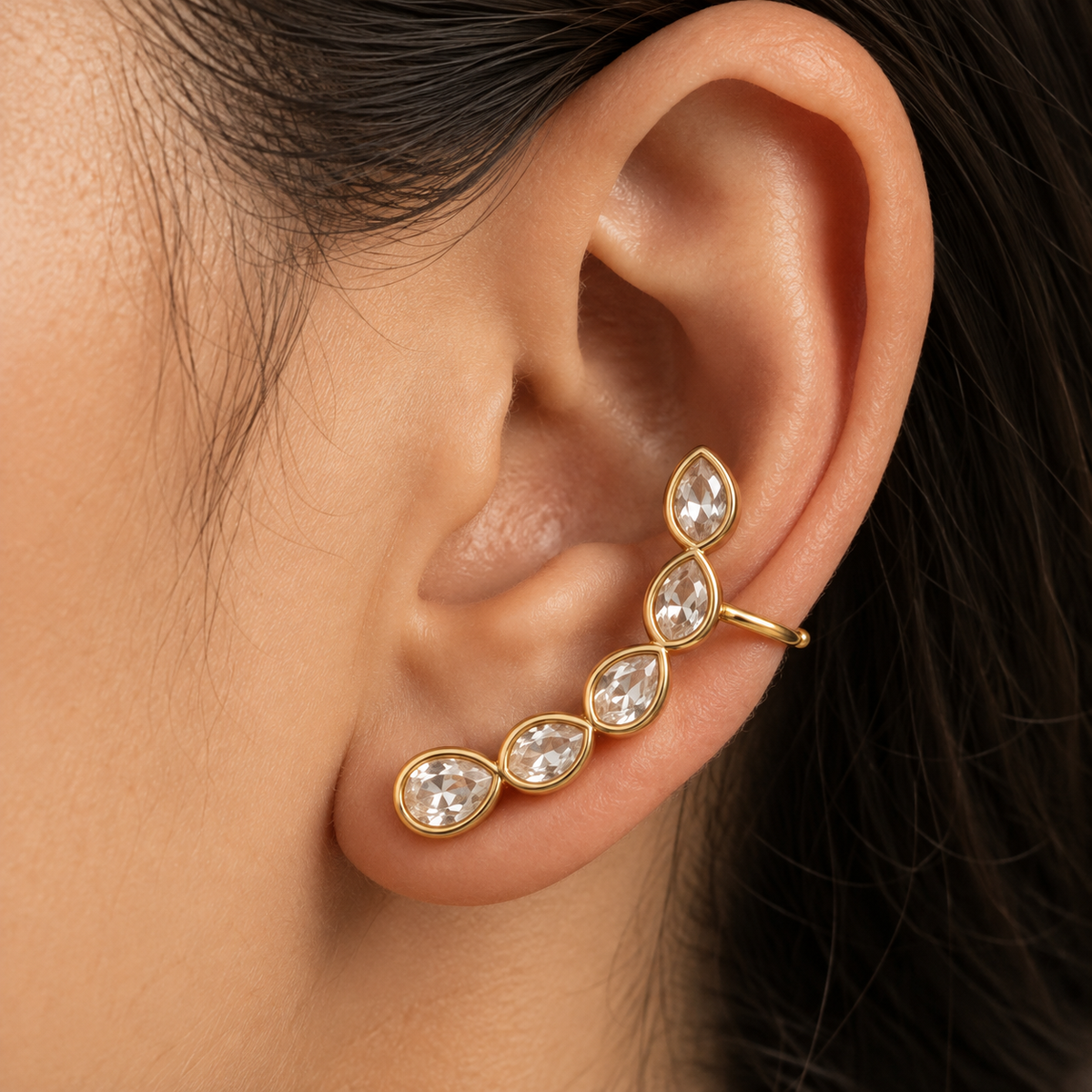 A close-up of a person's ear wearing a gold ear cuff with five teardrop-shaped clear gemstones.