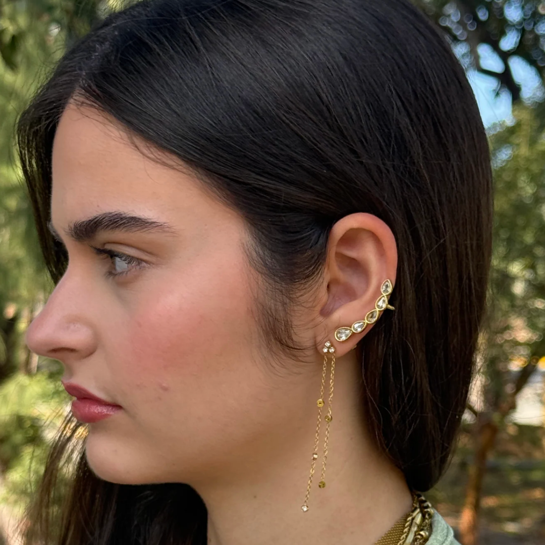 A close-up side profile of a woman with dark hair wearing a gold ear cuff with small clear stones and dangling gold chain earrings, set against a blurred outdoor background of green trees.