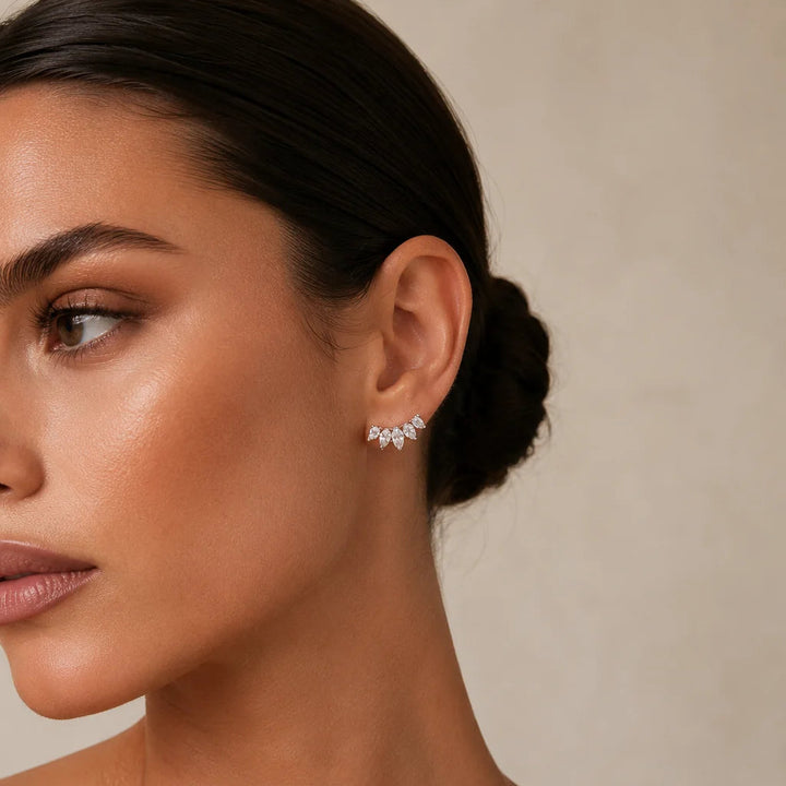 A close-up of a woman wearing a small silver crescent-shaped stud earring with clear gemstones, set against a neutral beige background.