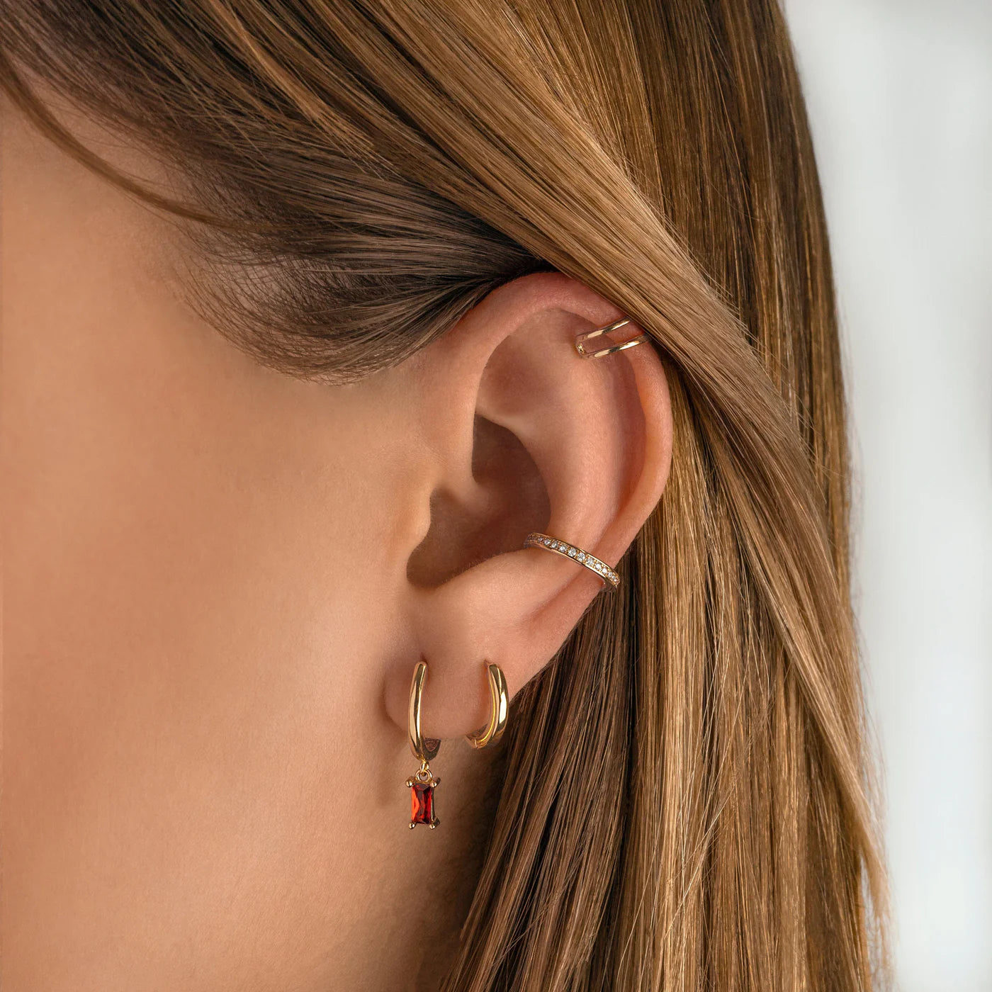 Close-up of an ear wearing gold hoop earrings with a red gemstone and a small diamond ring.