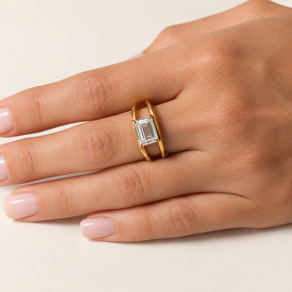 A gold ring with a rectangular emerald-cut clear gemstone, shown worn on a hand with pale pink manicured nails against a light beige background.