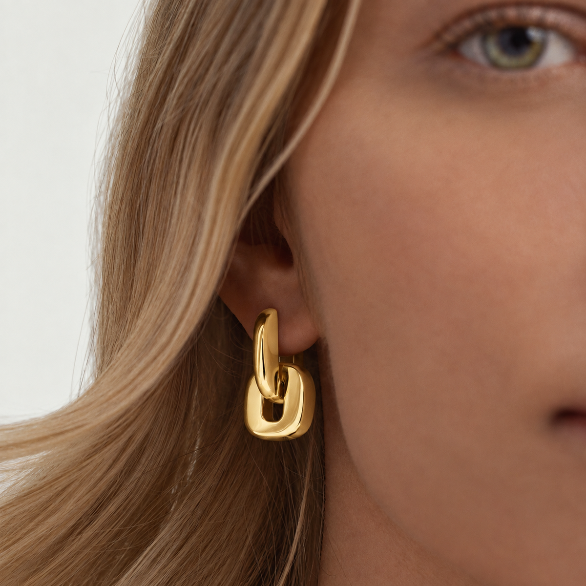 A close-up of a model's ear wearing a polished gold huggie hoop earring with a thick interlocking link design.
