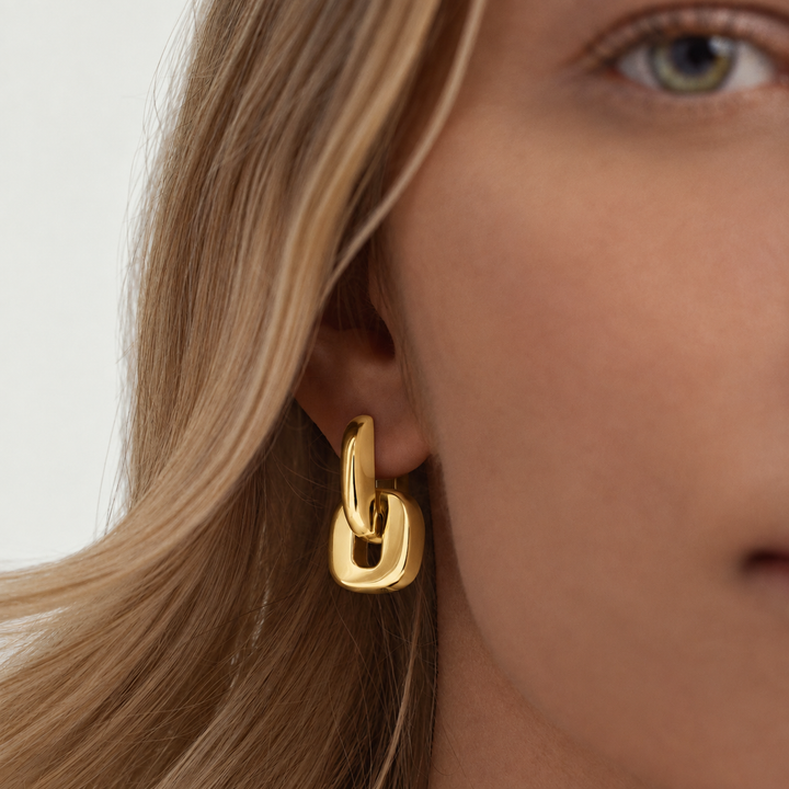 A close-up of a model's ear wearing a polished gold huggie hoop earring with a thick interlocking link design.
