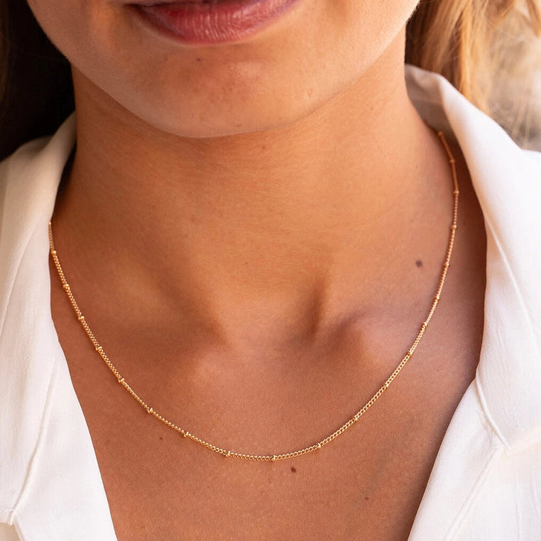 A close-up of a woman's neck and upper chest wearing a delicate gold chain necklace with small bead-like links, paired with a white collared shirt.