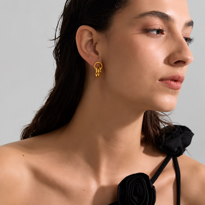 A close-up side profile of a woman wearing a small gold hoop earring with three dangling textured loops. She has dark hair and is wearing a black floral halter top against a plain light gray background.
