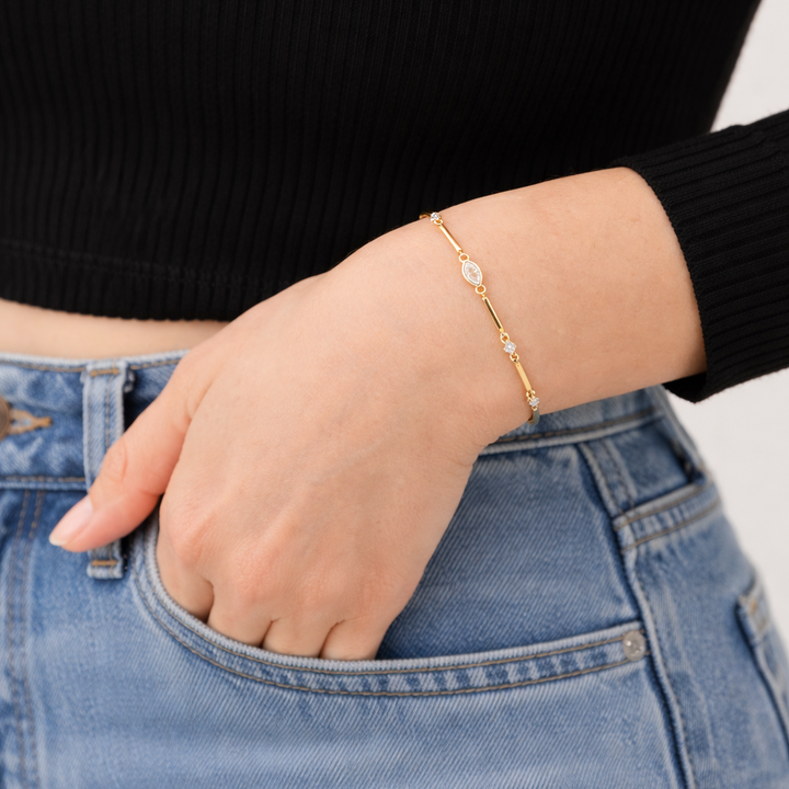 Hand wearing a gold bracelet with small pearls, against a black top and blue jeans background.