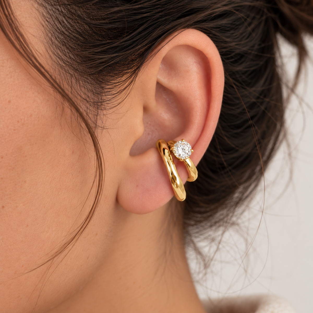 A close-up of a person's ear wearing a pair of gold huggie hoop earrings, one of which features a small round clear gemstone, against a soft light background.