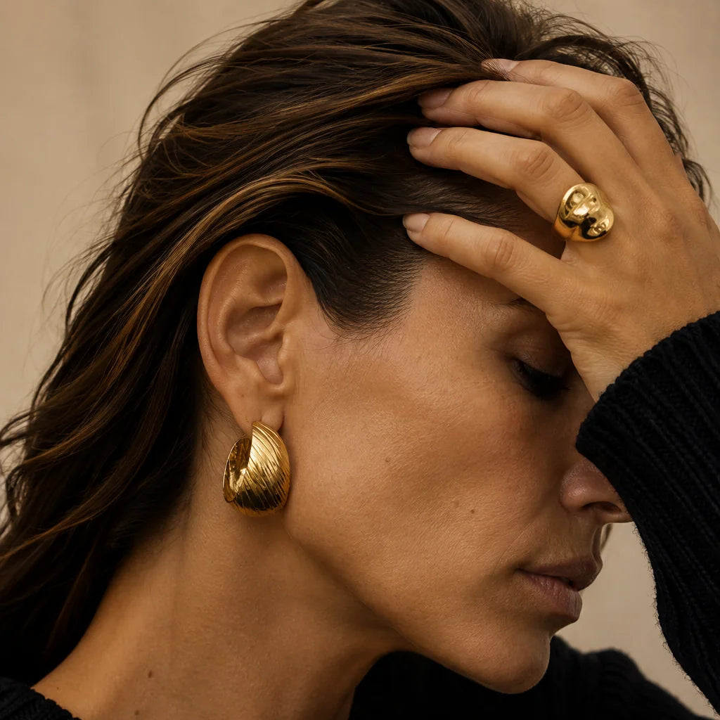 A close-up lifestyle shot of a woman wearing textured gold hoop earrings and a matching gold ring, dressed in a black knit sweater against a neutral beige background.
