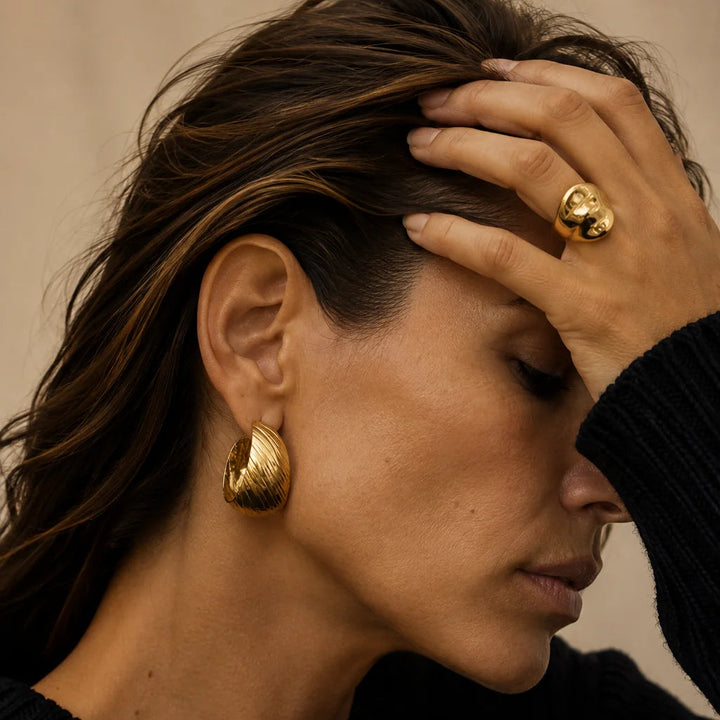 A close-up lifestyle shot of a woman wearing textured gold hoop earrings and a matching gold ring, dressed in a black knit sweater against a neutral beige background.