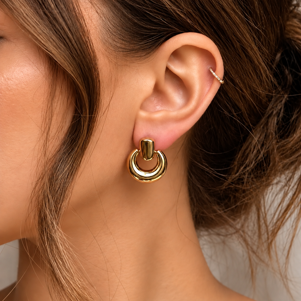 A close-up of a woman's ear wearing a polished gold hoop earring with a smaller gold ring dangling from it, alongside a delicate gold ear cuff.