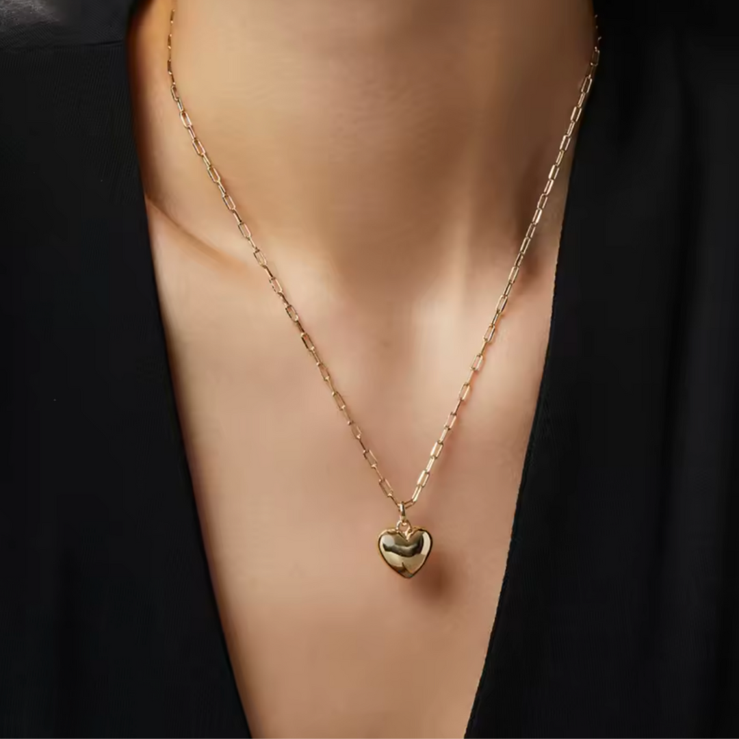A close-up of a person's neck and chest wearing a delicate gold chain necklace with a small polished heart pendant, styled with a black top.