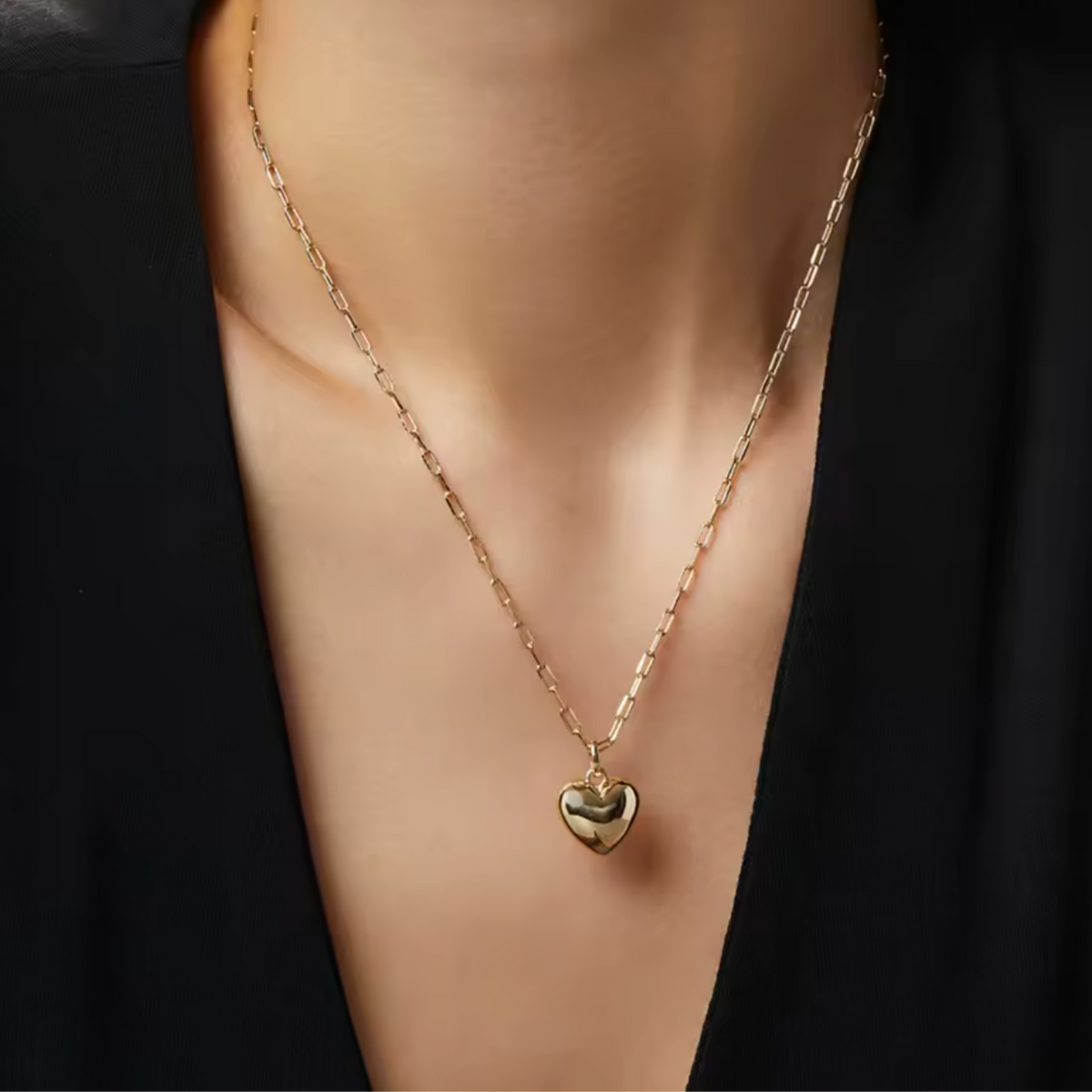 A close-up of a person's neck and chest wearing a delicate gold chain necklace with a small polished heart pendant, styled with a black top.