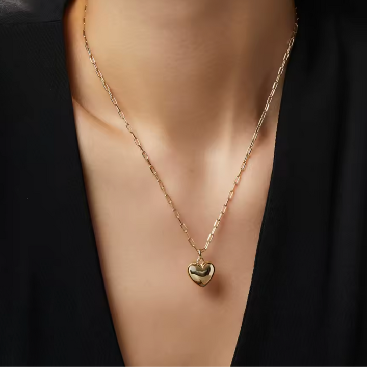 A close-up of a person's neck and chest wearing a delicate gold chain necklace with a small polished heart pendant, styled with a black top.