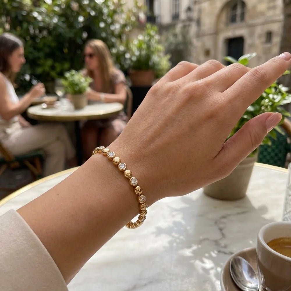 A gold beaded bracelet with clear rhinestone accents worn on a woman's wrist, photographed in an outdoor cafe setting with blurred patrons in the background.