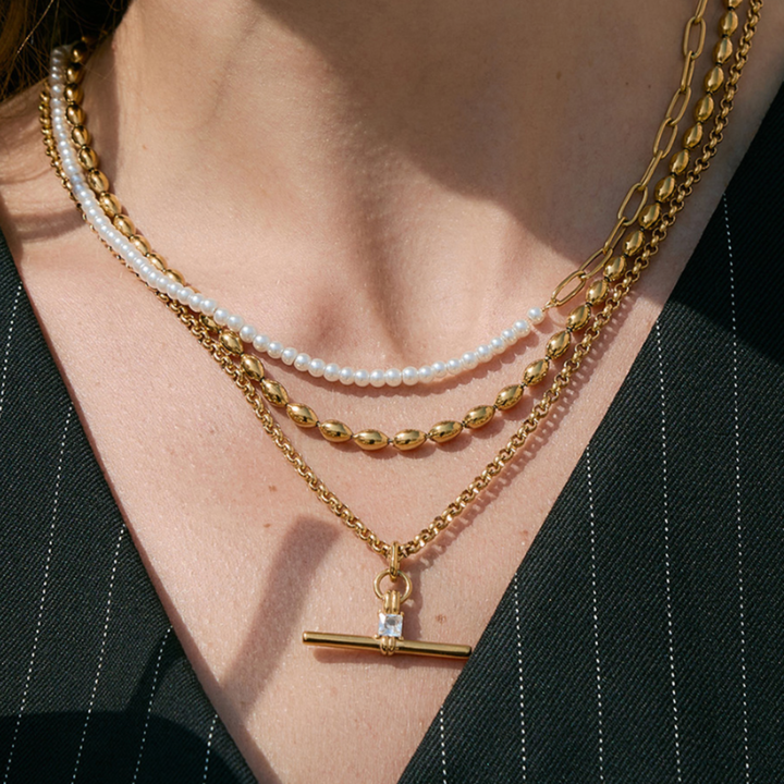 A close-up of a person's neck and chest wearing a layered necklace set. The set includes a gold chain with a bar pendant, a gold beaded chain, a white pearl strand, and a gold paperclip chain, styled with a black pinstripe top.