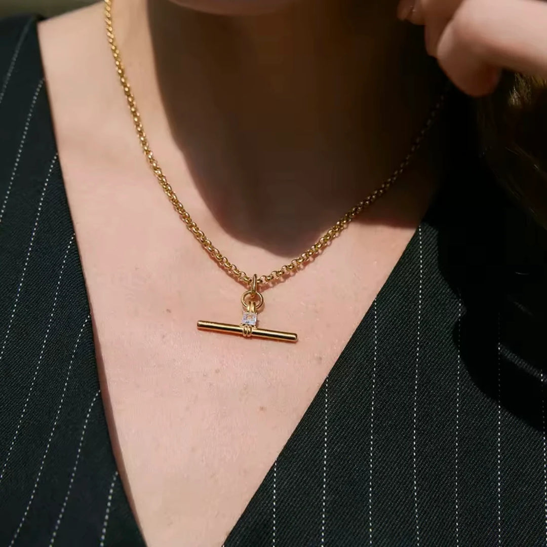 A close-up of a woman wearing a gold chain necklace with a toggle bar pendant featuring a small clear gemstone, styled with a black pinstripe top.