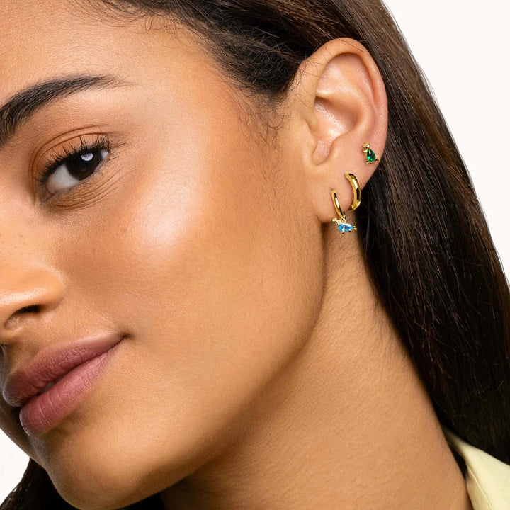 Close-up of a woman's ear wearing three small gold hoop and stud earrings, including one with a green stone and one with a light blue stone, against a clean white background.