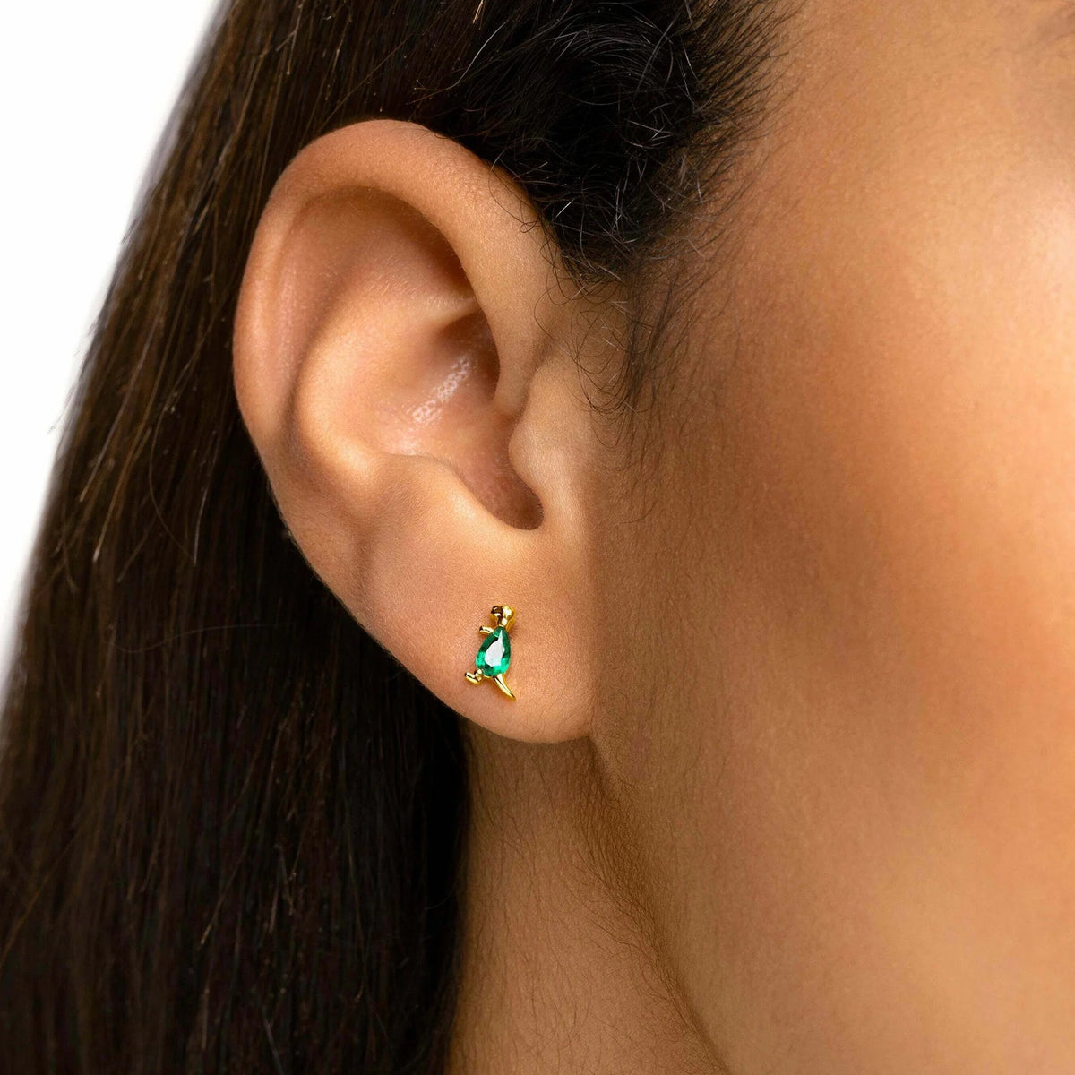 A close-up of a person's ear wearing a small gold earring with a green teardrop-shaped stone.