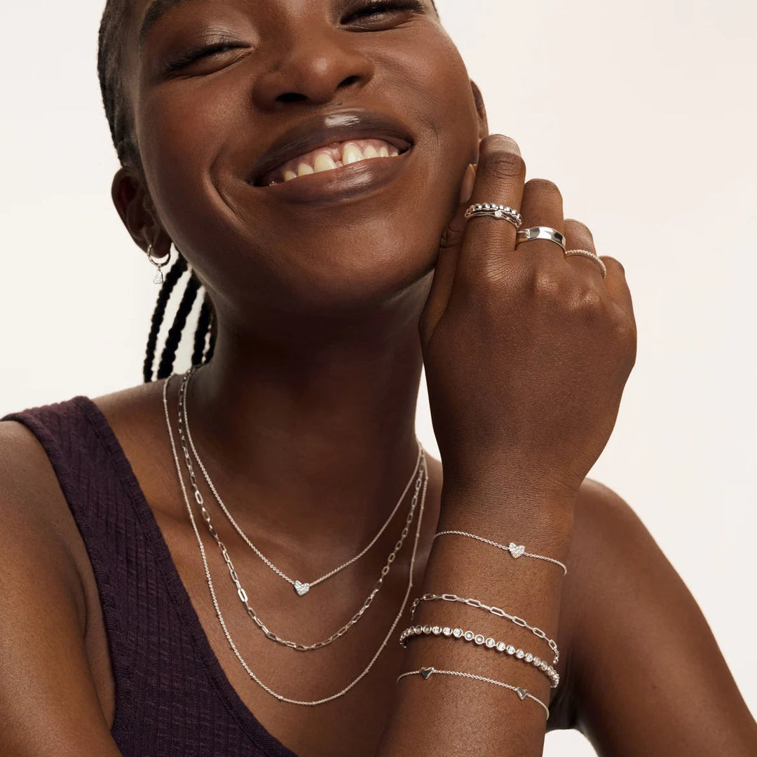 Woman wearing multiple silver jewelry pieces including necklaces, bracelets, and rings on a neutral background