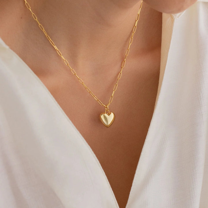 A close-up of a person wearing a delicate gold chain necklace with a small polished heart-shaped pendant, styled with a white V-neck top.