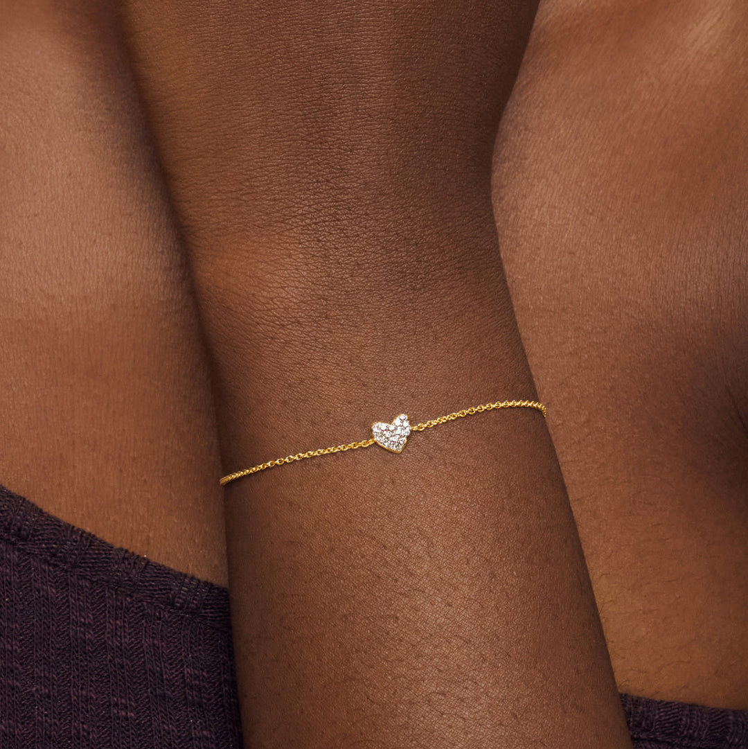 Gold bracelet with a heart-shaped charm on a person's wrist against a brown background