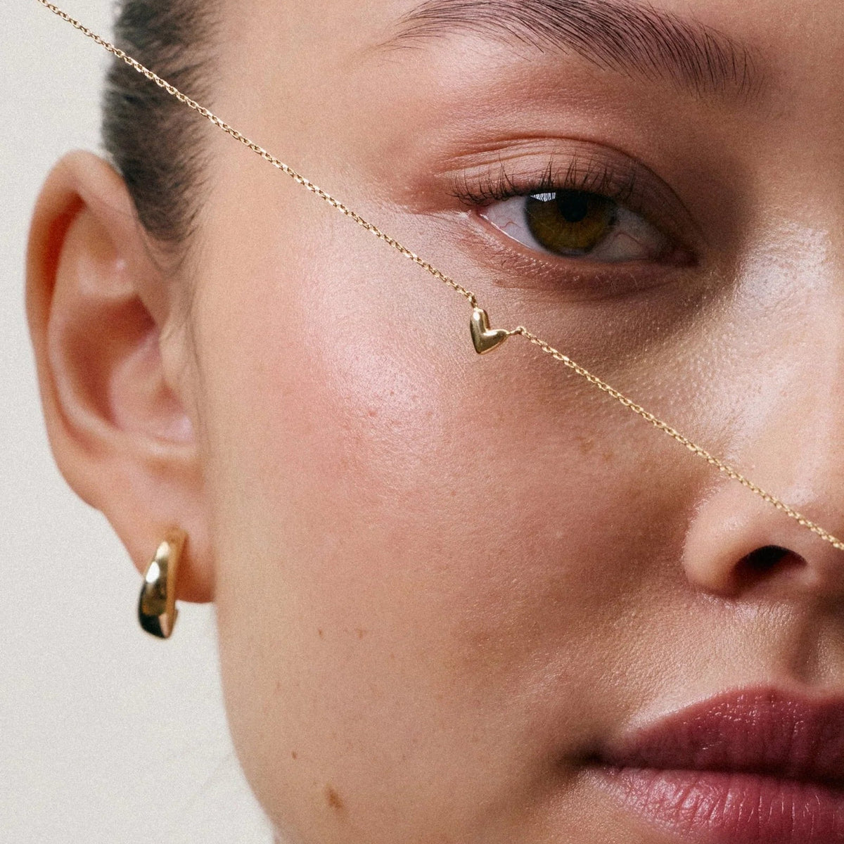 Close-up of a model wearing a delicate gold chain necklace draped across her face and a small gold hoop earring, against a soft beige background.