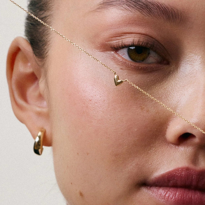 Close-up of a model wearing a delicate gold chain necklace draped across her face and a small gold hoop earring, against a soft beige background.