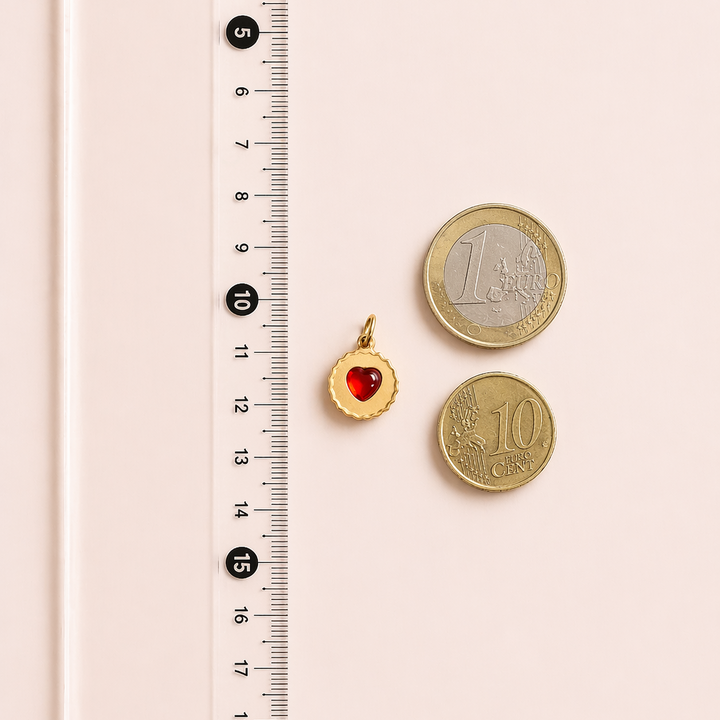 Small gold pendant with a red heart next to two gold coins on a pink surface with a ruler.