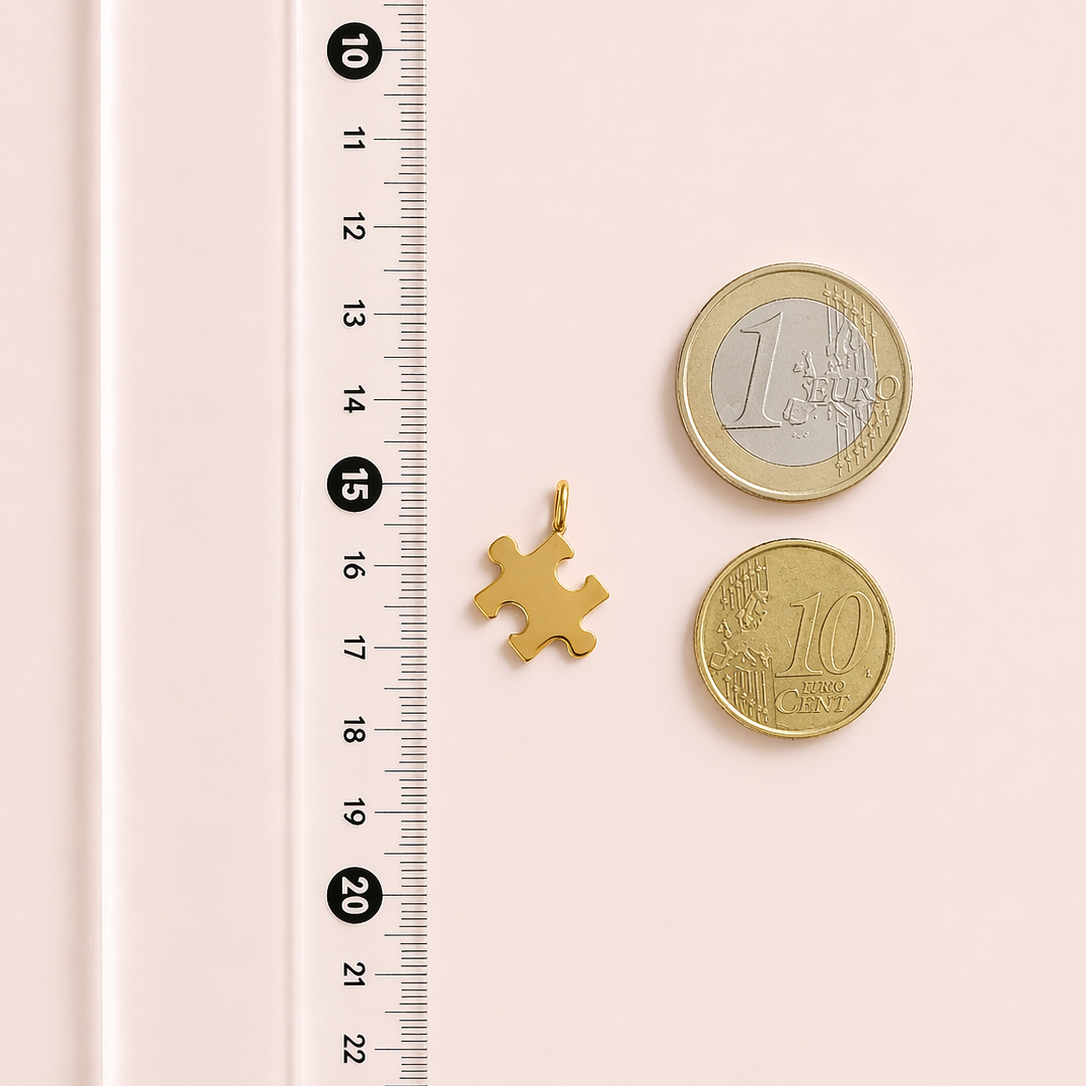 Gold puzzle piece charm next to two gold coins on a pink background with a ruler for scale.