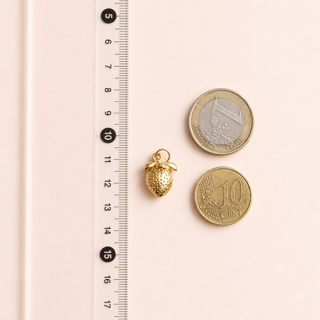 Gold strawberry charm next to two coins on a pink surface with a ruler for scale.